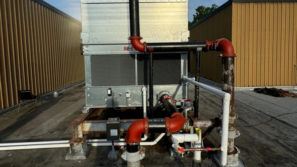 An HVAC unit with red and black pipes on a flat roof with brown buildings and a blue sky in the background.